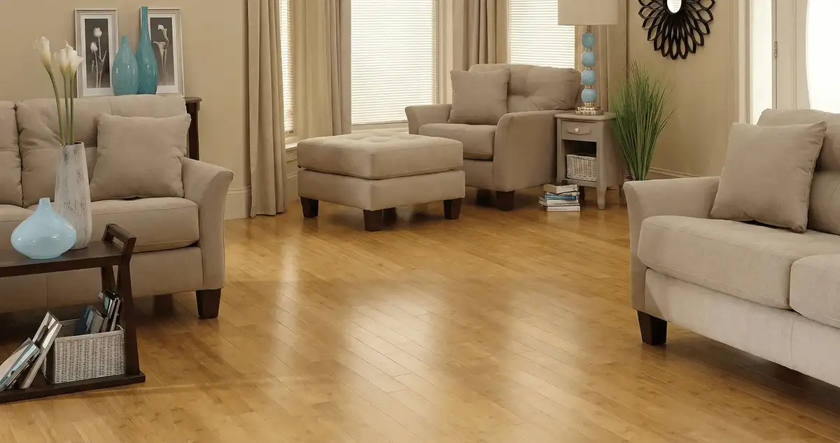 A living room with professionally installed bamboo flooring, beige sofas, a matching ottoman, a side table with a blue lamp, vases, framed pictures, and large windows with blinds and curtains in Philadelphia, PA